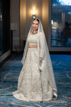  Ami in a white and silver embroidered lehenga with a sheer veil, smiling confidently indoors before her wedding ceremony.