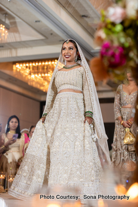 The bride wearing an elegant white lehenga with intricate designs and matching jewelry, standing in a beautifully lit hallway.