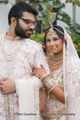  The bride and groom smiling together in pastel pink and cream wedding attire with an elegant backdrop. 