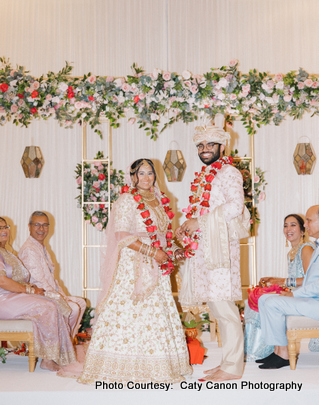 The bride and groom smiling together in pastel pink and cream wedding attire with an elegant backdrop.