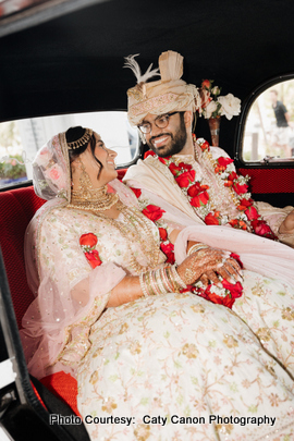 The bride and groom smiling together in pastel pink and cream wedding attire with an elegant backdrop.